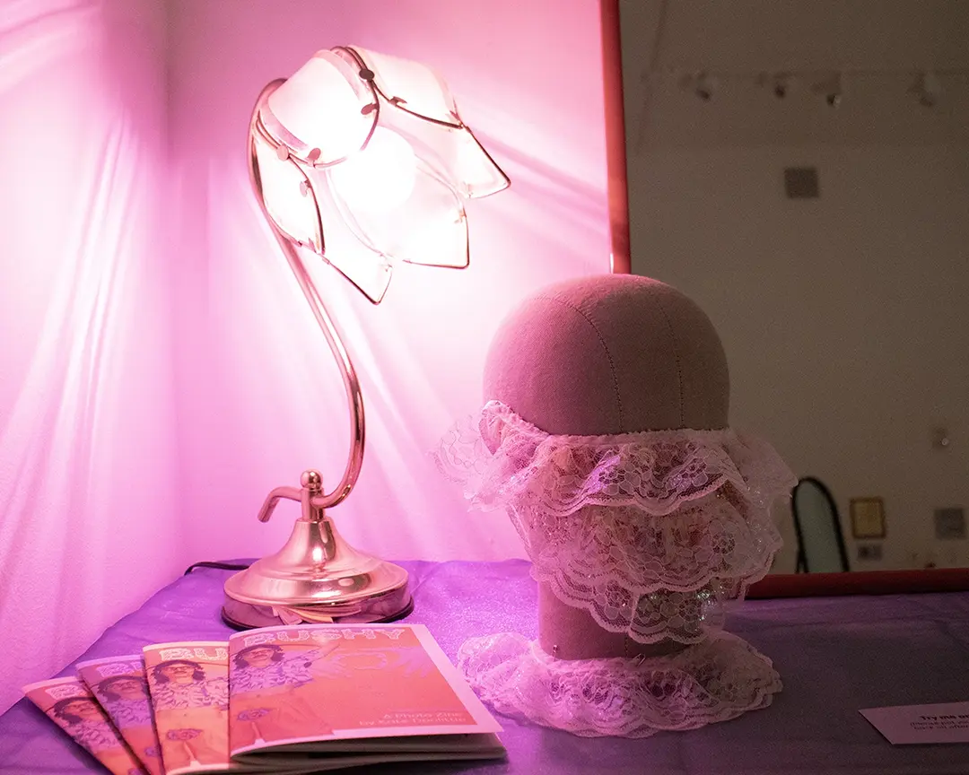 Mannequin head wearing a lace beard with a lace collar around the base. It's on 
                    a table with a sheer iridescent blue fabric over it, sitting in front of a mirror. 
                    Behind the head is a floral shaped lamp with a pink light casting patterns on the wall. 
                    There's also a stack of zines on the table.