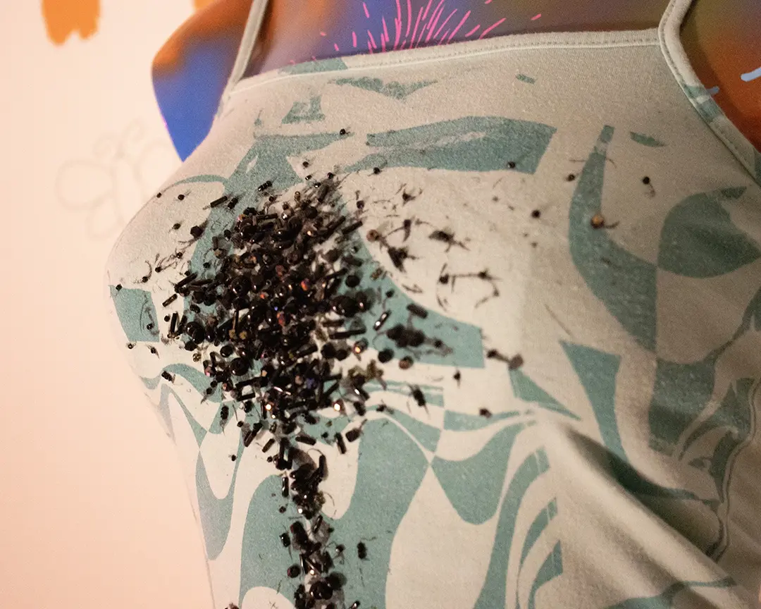 Low-light close up of a teal tank top on a colorful mannequin bust with black beaded chest hair 
                    on it. The beads making up the chest hair are very small and some are iridescent. There's a pink reflection 
                    from an off camera lamp in some of the beads.
