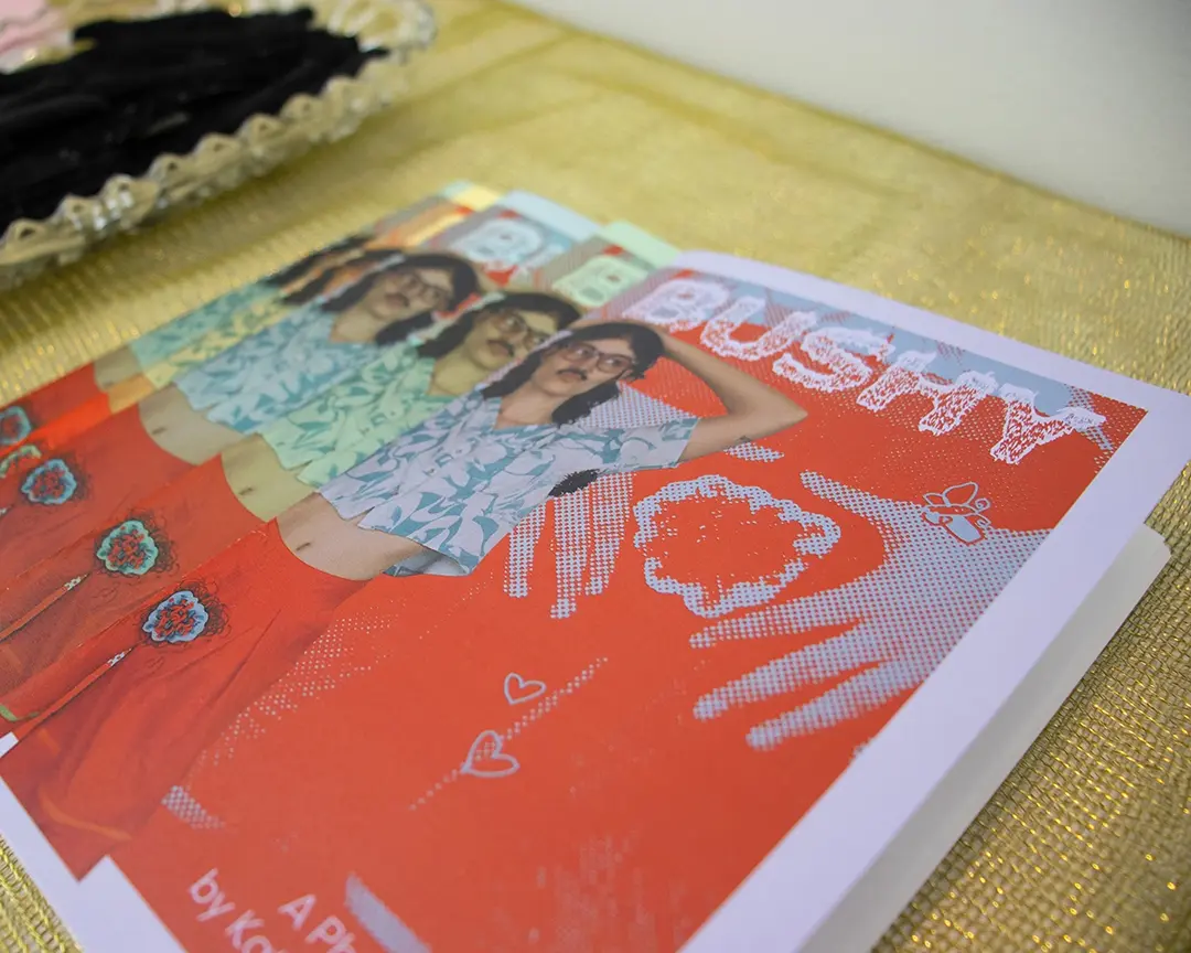 Close up of a stack of zines spread out on a pedestal. The pedestal is covered in a 
                    sheer, iridescent green fabric. Kate is on the cover of the zine wearing body hair clothes 
                    and accessories. The zine is titled the same as the show.