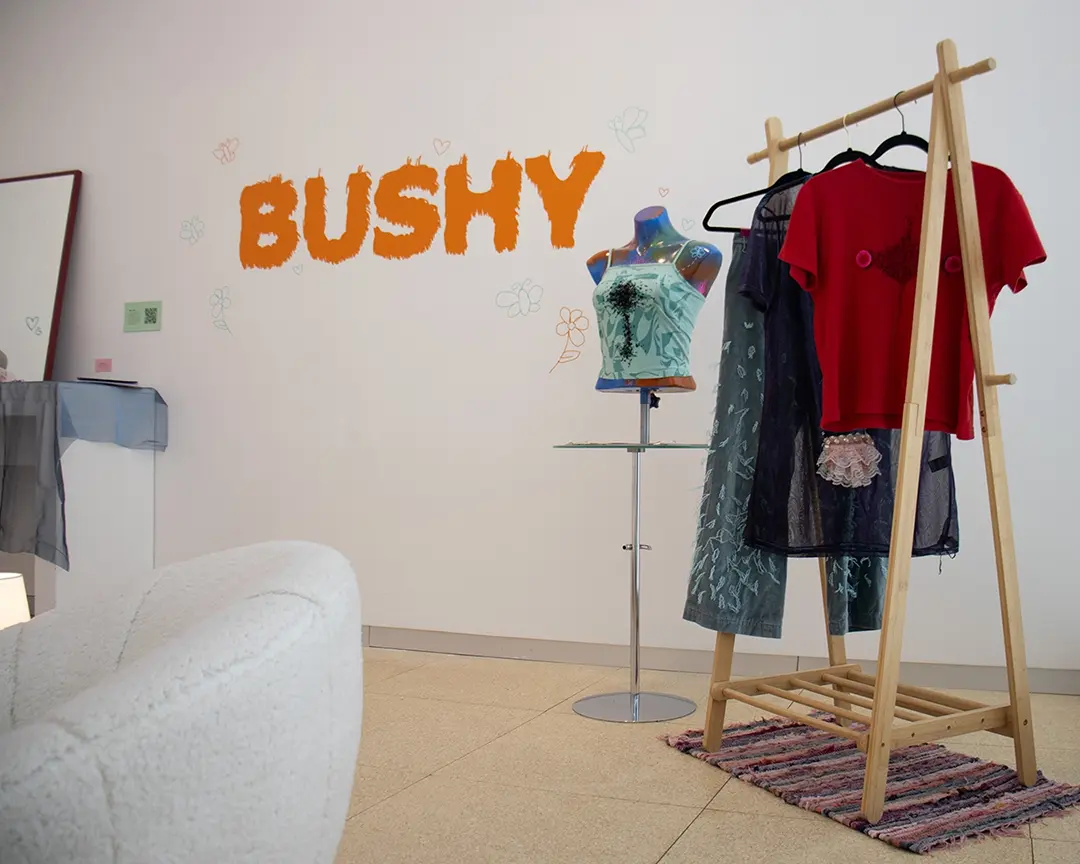 The back right corner of the gallery. There's a clothing rack with leg hair pants, 
                    a dress with a lace bush on it, and a shirt with chest hair and fake nipples on it. To the left 
                    of the rack is a small table with a mannequin bust on it wearing a black beaded chest hair shirt. 
                    On the wall is the title vinyl that says: BUSHY. There's a white fuzzy chair in the foreground 
                    partially off camera and a vanity setup in the back, also partially off camera.