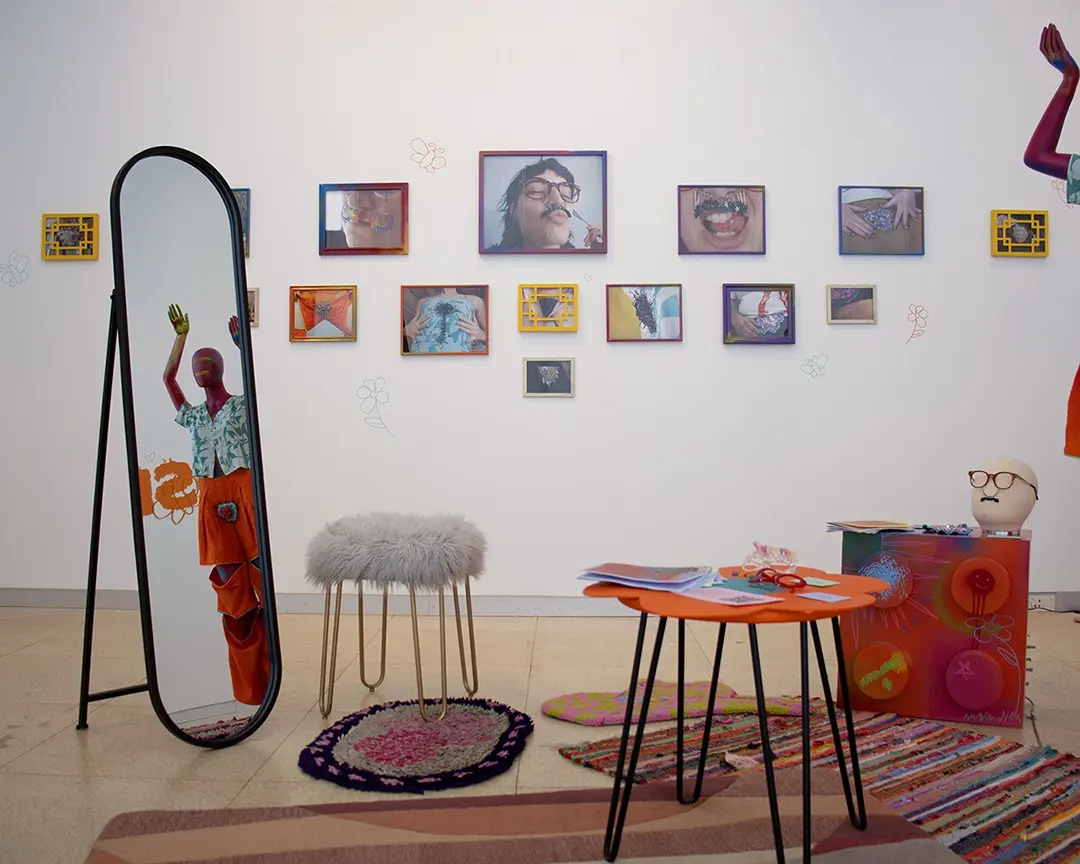 View of the left gallery wall which has many photos of people including Kate wearing 
                    body hair clothes and accessories. The photos are in colofrul frames of various sizes. In the 
                    foreground to the left is a floor-length standing mirror and a fuzzy stool. In the right-middle is an 
                    orange table shaped like a flower with zines and accessories on it. To the right is a table shaped like 
                    a giant lego with a mannequin head on it, and to the right of that, mostly off camera, is a full body 
                    mannequin with it's arm in the air.