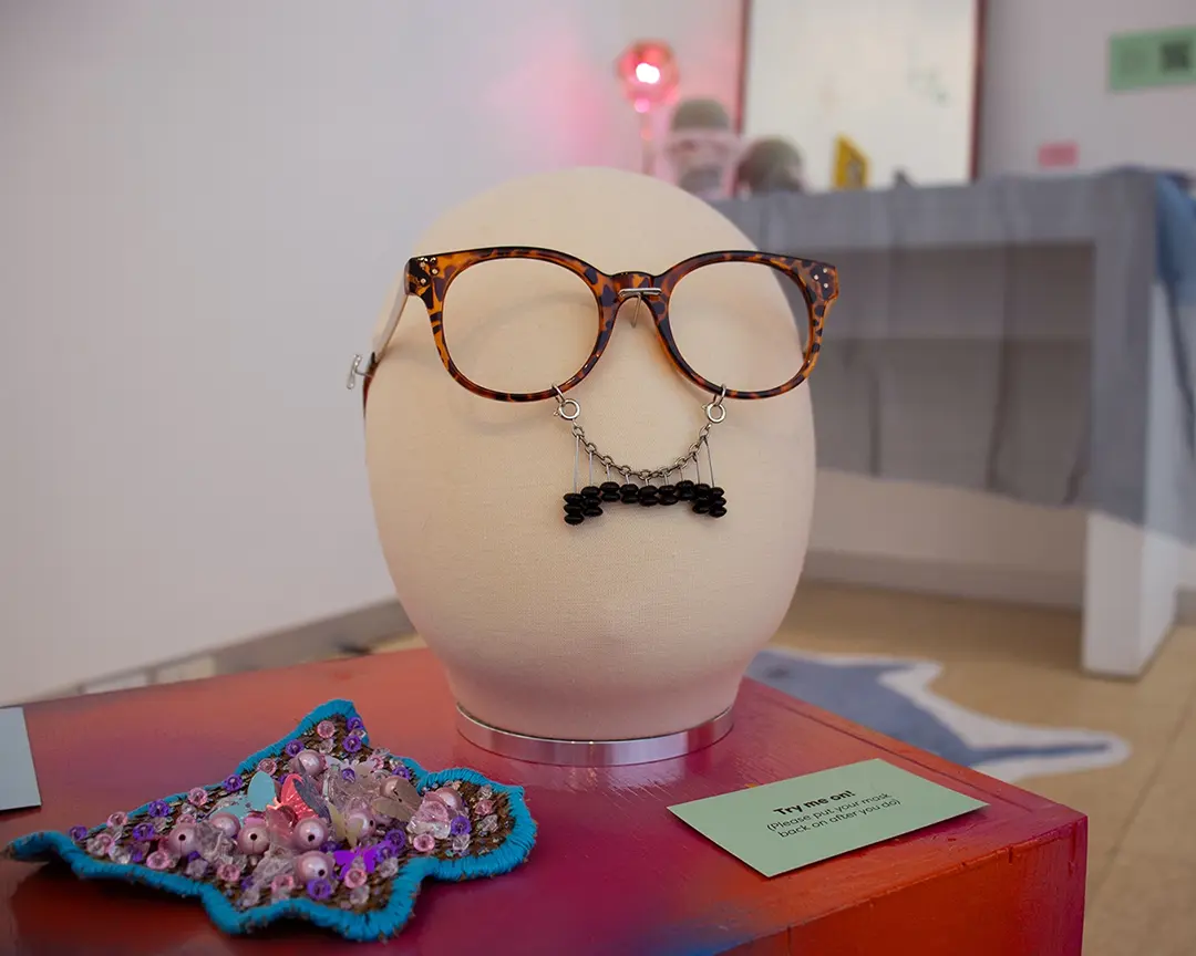 Close up of a mannequin head on a low table. It's wearing tortoise shell glasses frames with 
                    a black beaded mustache hanging from them. To the left is a pink, blue, and purple beaded bush in 
                    the shape of a butterfly made with butterfly shaped beads and sequins. Behind the head, out of focus, 
                    is a vanity set up.