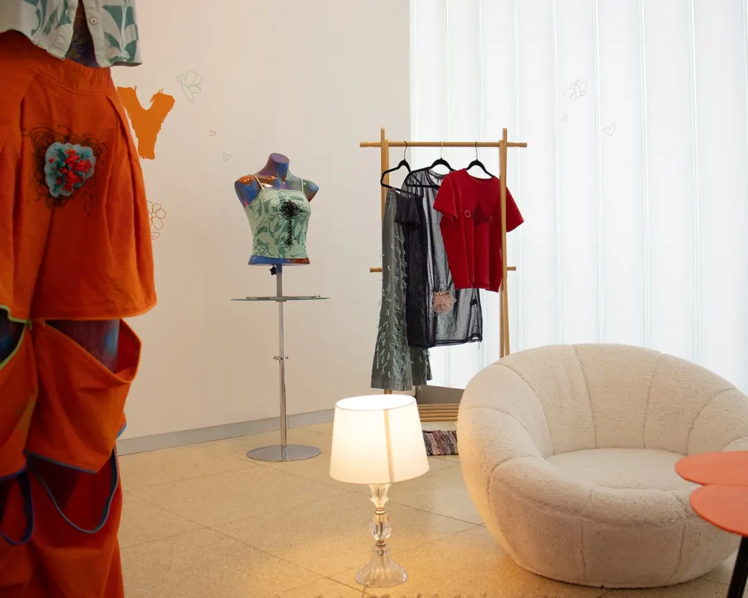 Another view of the back right corner of the gallery with a clothing rack and mannequin bust.
                    In the foreground to the left, partially off camera, is a full body mannequin wearing orange convertable 
                    pants with the zippers partially unzipped. In the foreground is a white fuzzy chair and a lamp giving 
                    warm soft lighting.