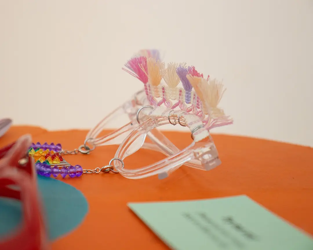 Close up of a pair of clear framed glasses on an orange flower table. They have a pink, purple, and yellow 
                    tassel trim attached to the top like a unibrow, and a rainbow beaded mustache hanging from the frames. There's 
                    a pair of red framed glasses out of focus in the foreground, slightly blocking the rainbow mustache.