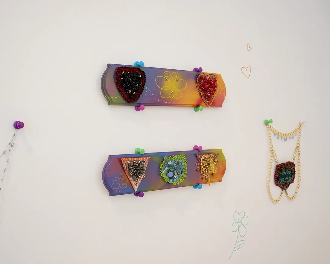 One wall of the gallery with two colorful boards hung up in the middle with large colorful 
                    push pins. On the top board are two mounted bushes with an empty space in the middle. 
                    The bottom board has three mounted bushes. To the left, out of frame, is a safety pin chain 
                    also hanging from a giant push pin. To the right is a harness, also hanging with giant push pins, 
                    that has a merkin in the shape of an archaic medusa head on it.