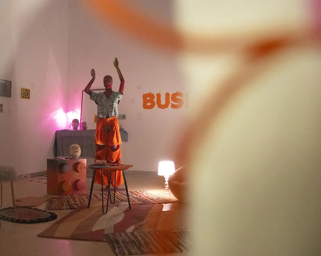 The gallery trhough a transparent shower curtain with orange doodles on it. There's a 
                    mannequin wearing body hair clothes, lots of rugs on the floor, and soft warm lamp lighting.