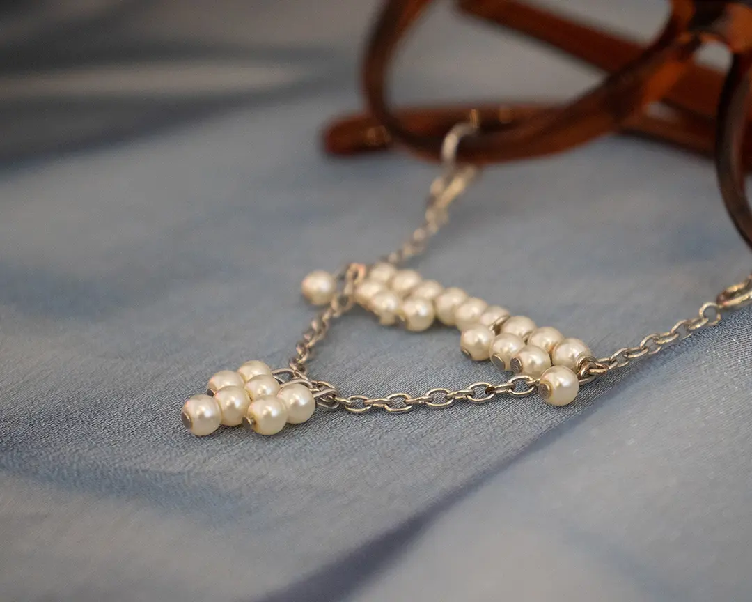 Close up of a pair of brown framed glasses with a beaded pearl mustache and soul patch 
                    hanging from the rims. It's sitting on blue iridescent fabric.