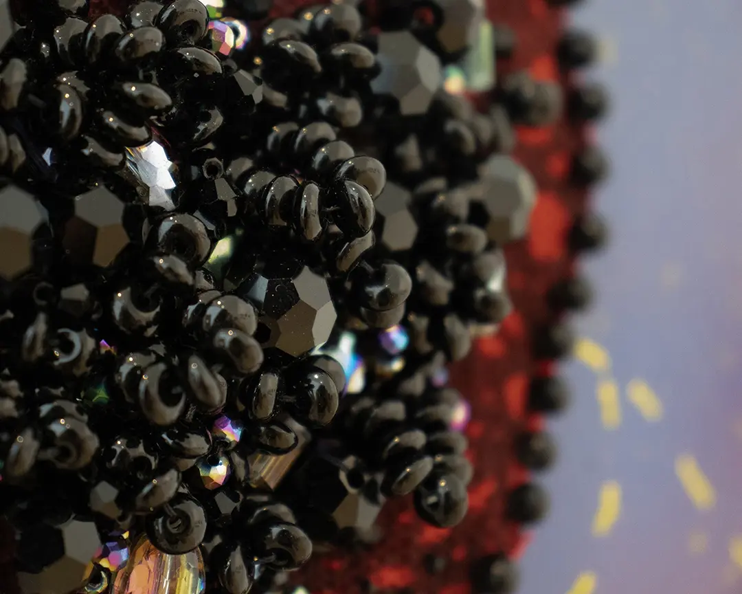 Close up of a beaded bush. It's made of smooth oval balck beads and iridescent 
                    beads forming a dense clump of pubic hair. It's sewn onto red fabric with a border of round 
                    black beads and it's mounted on a purple board.