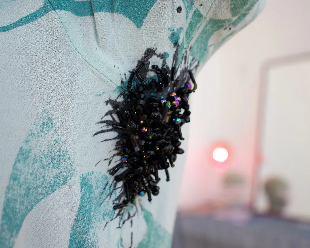 Close up of a shirt with beaded armpit hair on a mannequin. The black and iridescent beads 
                    are looped to look like thin strands of hair on the edges and more densly clustered hair in the 
                    middle. Out of focus in the background is a vanity set up.