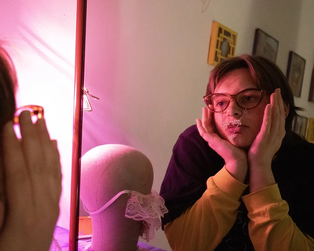 Nix looking in the mirror. The lighting is dark with a pink lamp casting a glow 
                    on their face. They're wearing brown framed glasses with a pearl mustache and soul patch 
                    hanging from them. Their chin is resting in their hands.