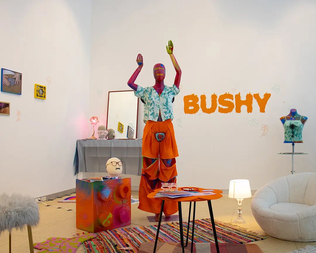Wide view of a gallery with a pink mannequin in the middle. There are two 
                    small tables with various accessories around it as well as a fuzzy chair, a fuzzy 
                    stool, and a lamp. There are layered rugs on the floor. In the back is a vanity set 
                    up with a mirror, pink lamp, and accessories. On the other side is a table with a 
                    mannequin bust wearing a beaded chest hair shirt. On the back wall is hairy vinyl 
                    lettering that says the show title: BUSHY.