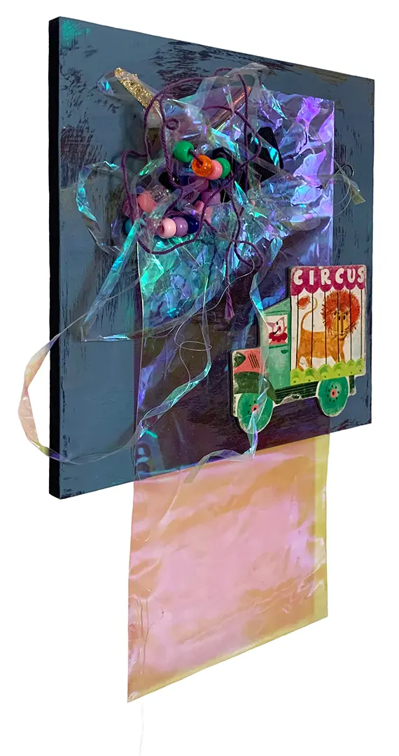 Left angle view of a three-dimensional collage on a painted board. It's made of miscellaneous 
                        trash, including strings of beads, plastic, and a circus cutout from a children's book.