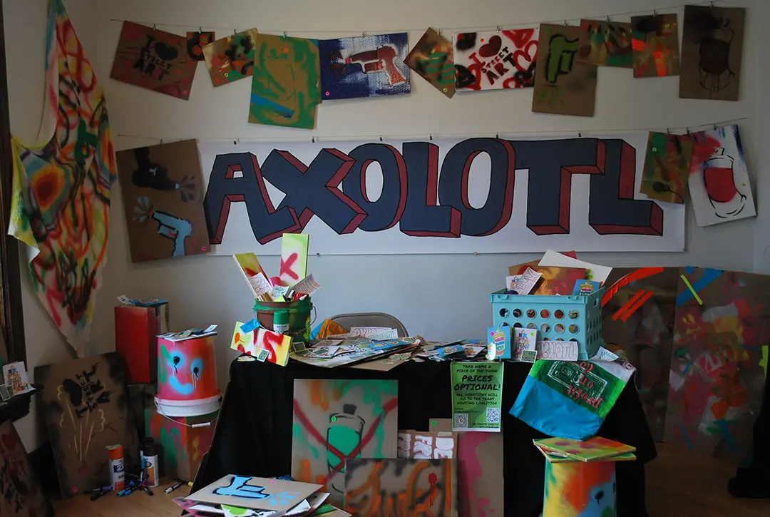 A table has spray painted cardboard signs, buckets, and wood panels, with zines, stickers,
                    and other materials scattered all around. The back wall has hanging cardboard signs and a large 
                    format sticker with the tag, 'AXOLOTL.'