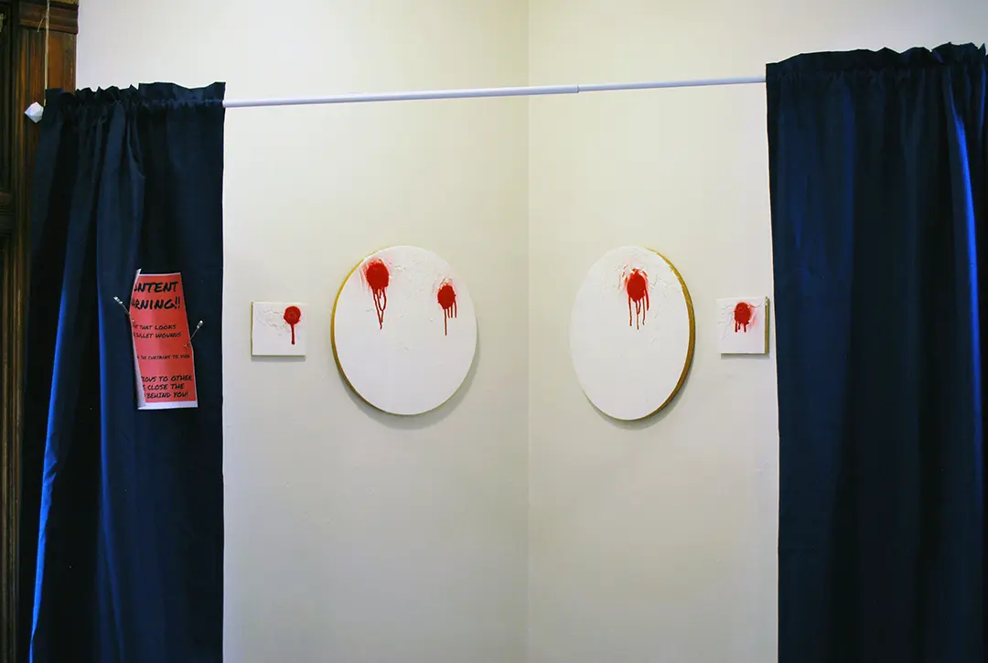 4 white paintings with red spray-paint that looks like bullet wounds. The paintings are behind 
                    a curtain with a content warning pinned to it, but the curtains have been pulled back for the photo.