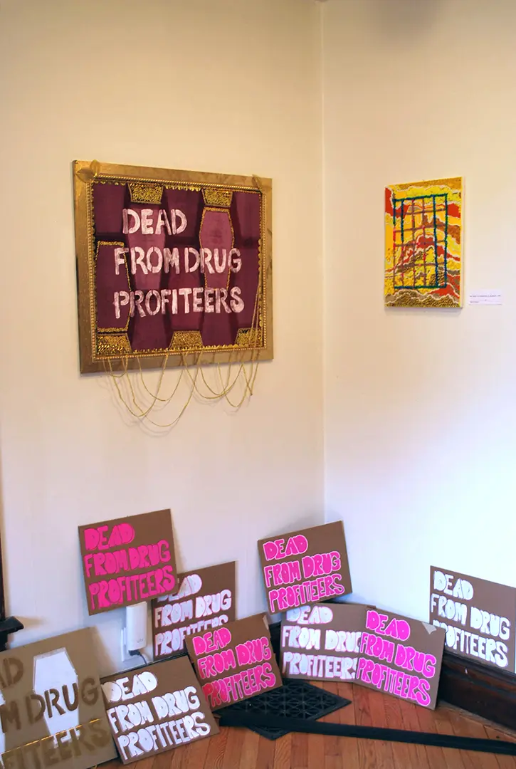 Velvet print in a gilded frame that says, 'DEAD FROM DRUG PROFITEERS.' 
                        Protest signs with the same phrase are on the ground. On the opposite wall is a bead painting.