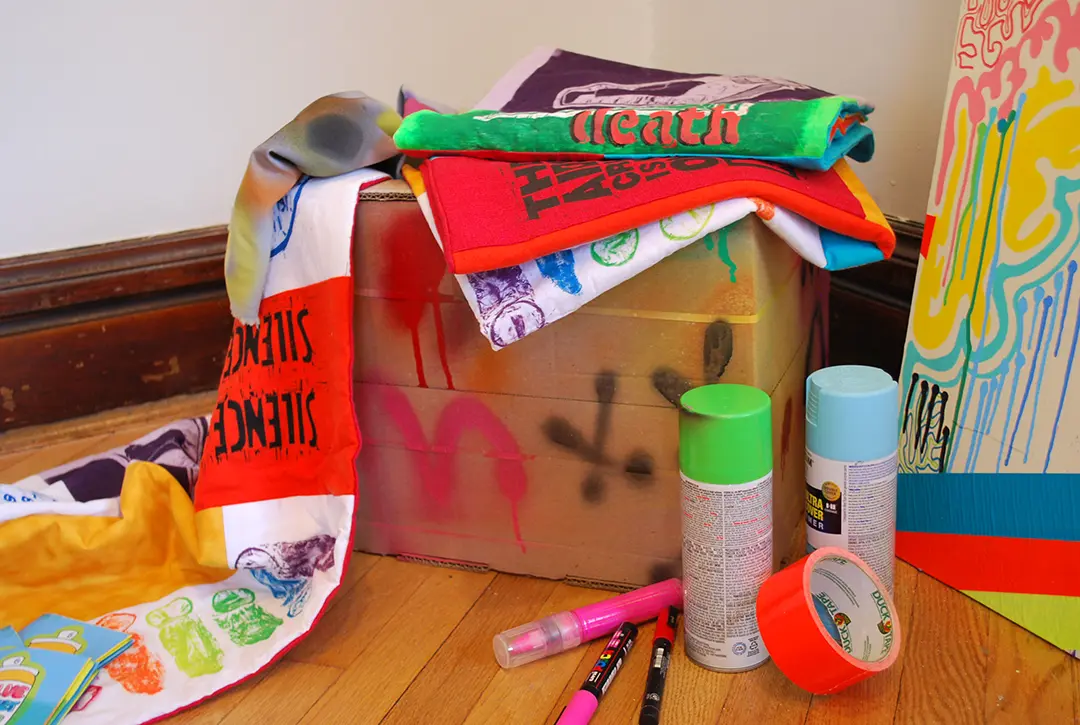 Close up of cardboard box and AIDS quilts. There are paint pens, spray paint, 
                    and duct tape on the ground.