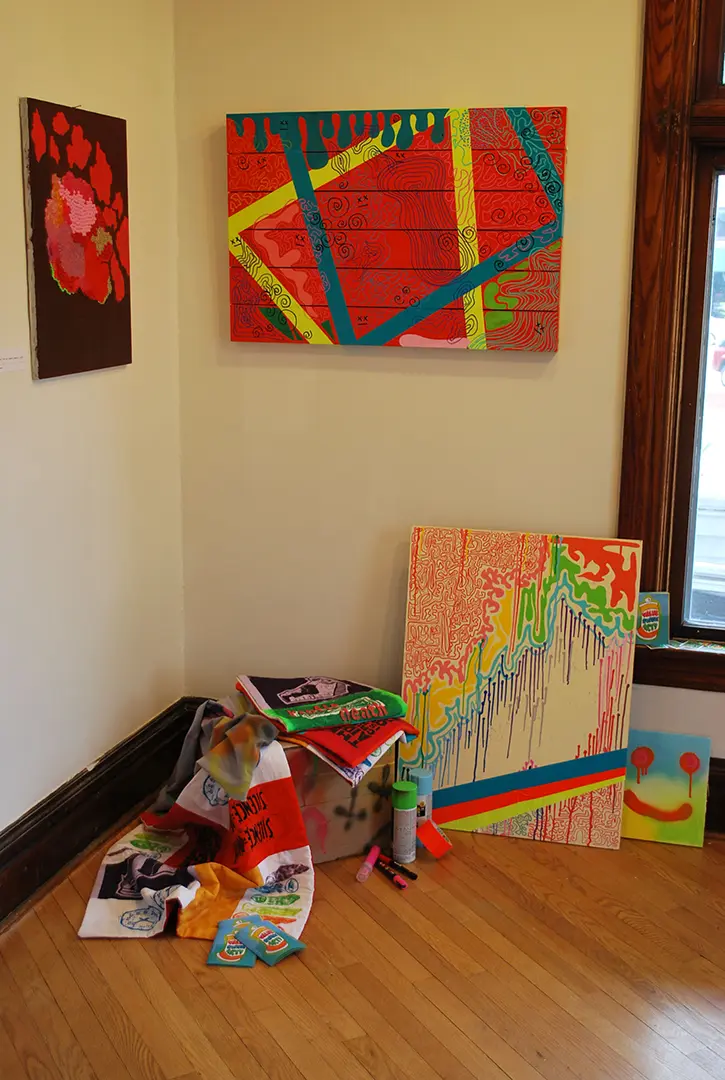 Bead painting on one wall and wooden painting on the other. On the floor is another 
                        wood painting, a cardboard box, and mini AIDS quilts.