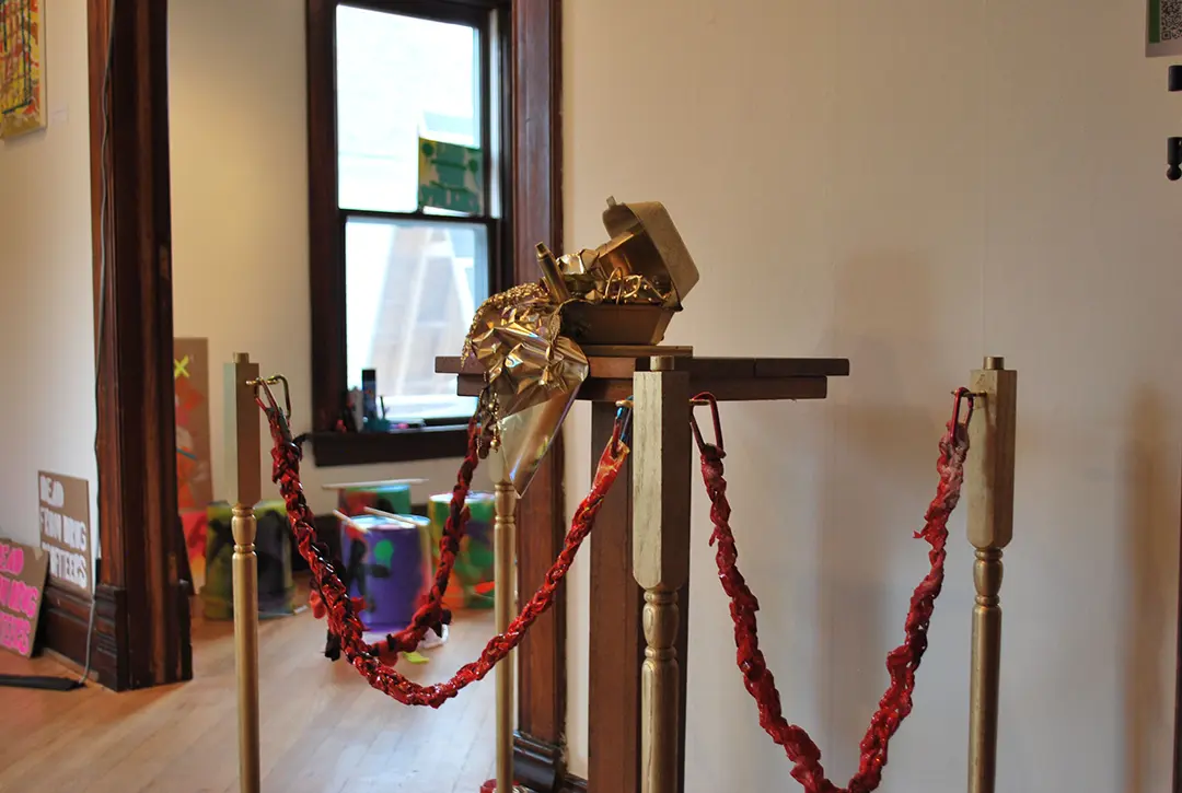 Wide angle of the treasure chest on the pedestal, surrounded by 'velvet' ropes made of trash 
                    and spray painted red. The ropes are clipped with carabiners to decorative wooden poles also spray painted gold.