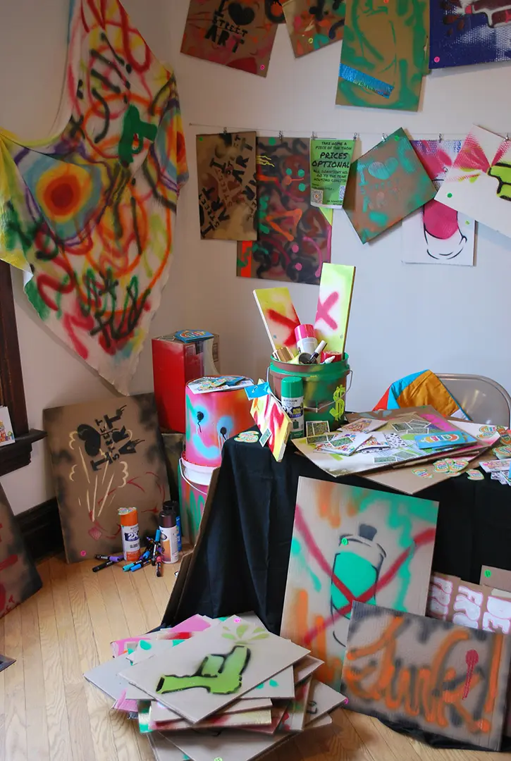 Corner to the left of the table filled with spray painted cardboard signs, 
                        buckets, and fabric. There are spray paint cans, paint pens, zines, and stickers 
                        scattered around.