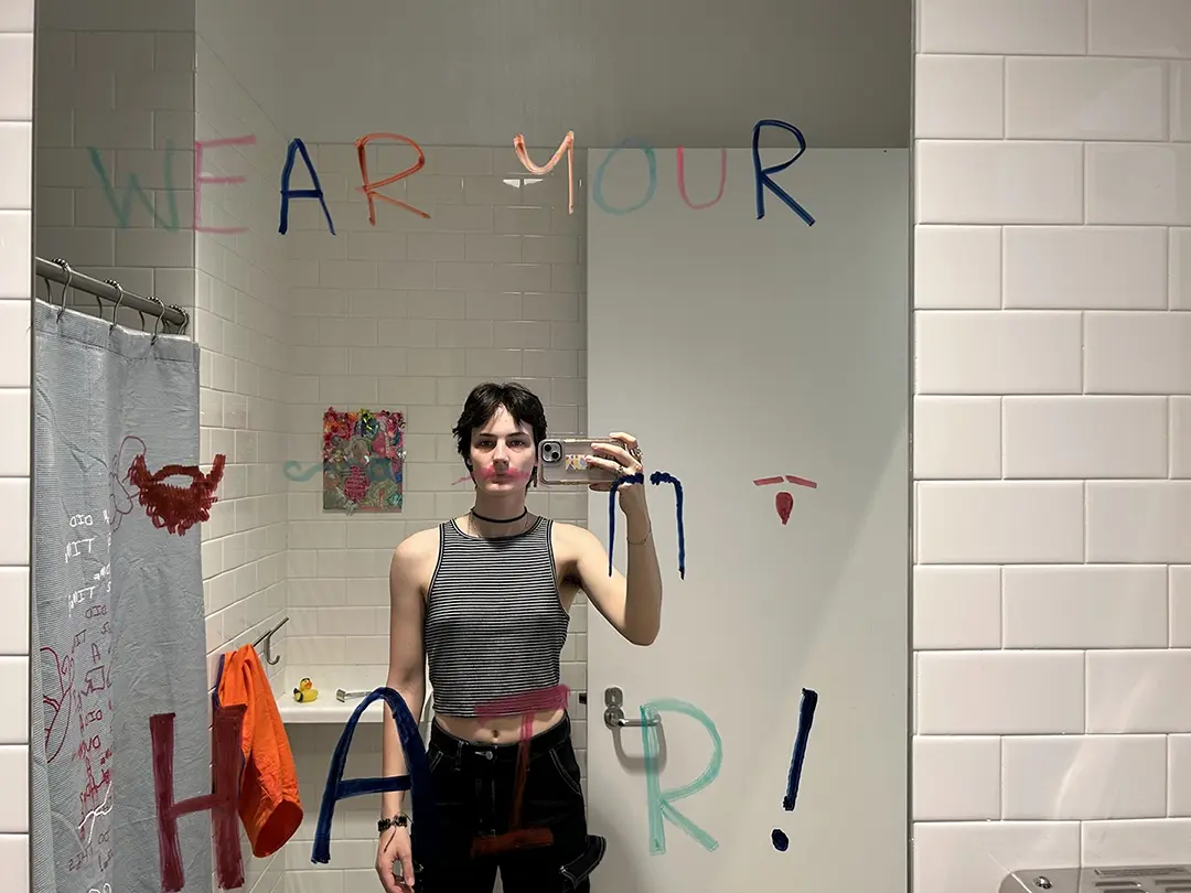 Mirror selfie of Kate. The mirror says "Wear your hair!" in expo markers and there are four facial 
                    hair variations for the viewer to stand in front of. Kate is standing in front of a pink mustache.