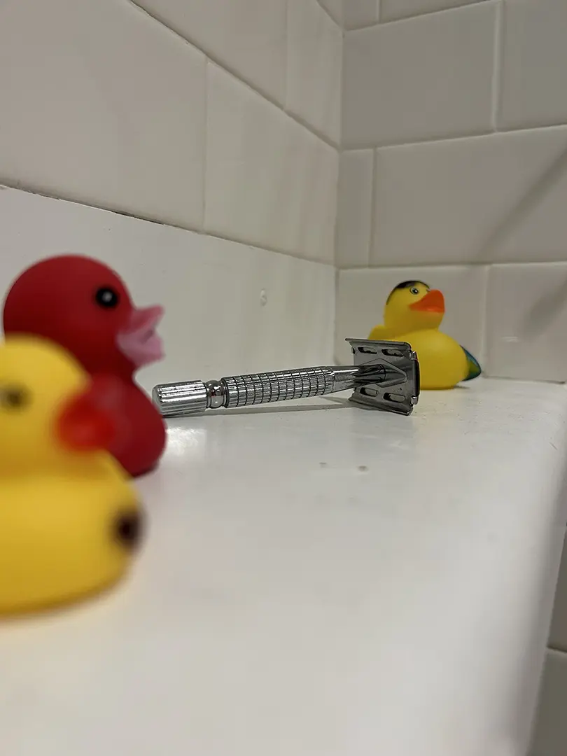 Close up of shelf with rubber ducks and safety razor. Two ducks are out of focus in the foreground, 
                        with the razor in focus in the middle.