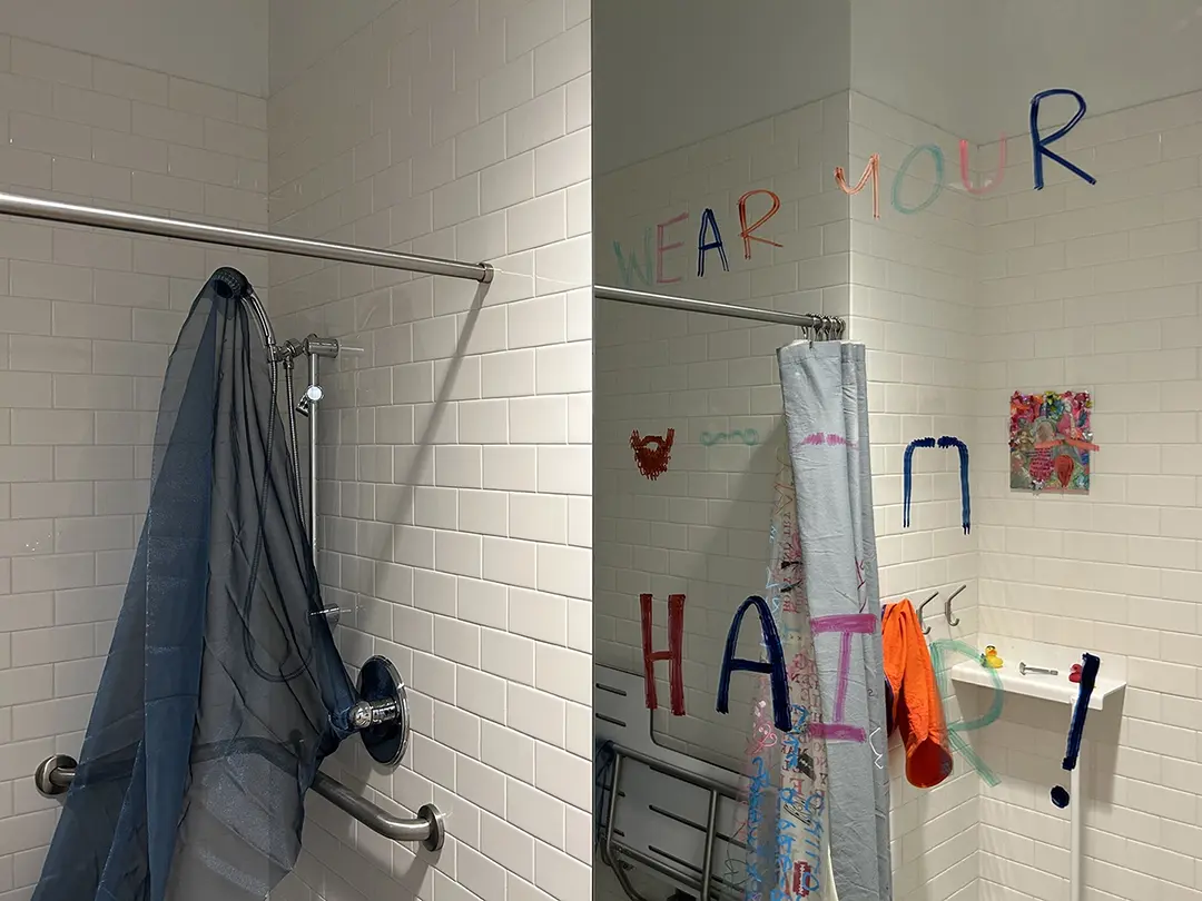 Close up of mirror and open shower. There are mustaches drawn on the mirror and text that reads: Wear your hair!
                    There's sheer blue fabric inside the shower.