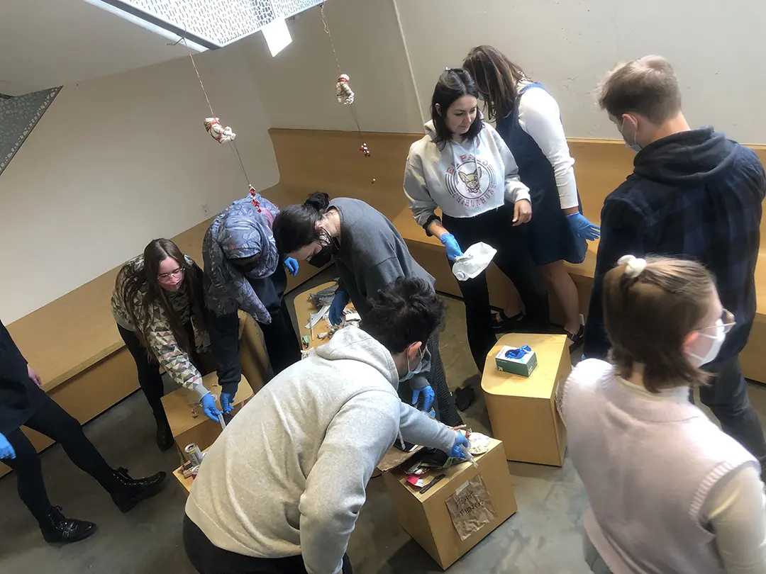 Still from a video of 'Sort Trash With Me!' Kate and their classmates are 
                    sorting trash that has been dumped on a pedestal in the Visual Arts Building.