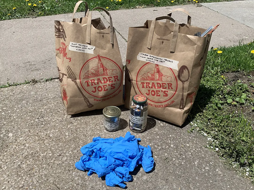 Two brown paper bags and two recycled jars full of categorized trash, 
                        and a pile of used blue nitrile gloves.