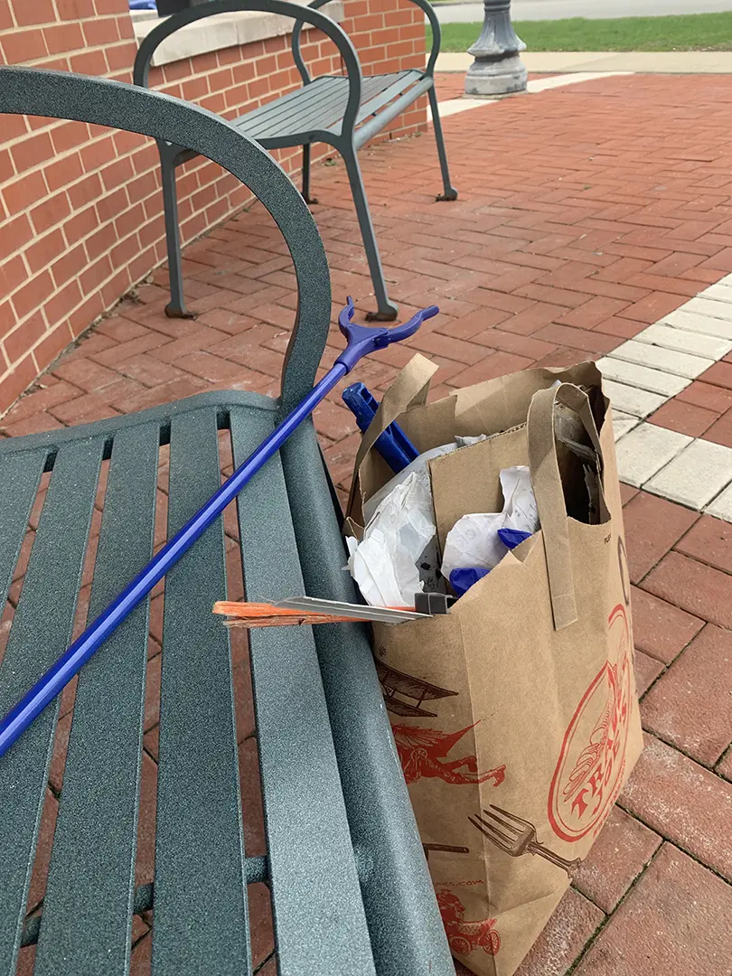 A brown paper bag filled with trash leaning against a bench. A blue reach 
                        extender is laying across the bench.