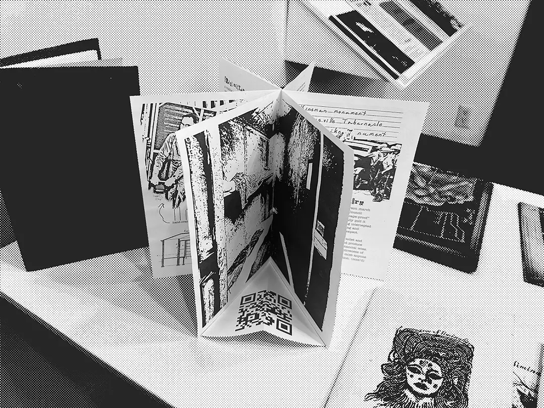 A zine standing on a pedestal. It's been bound rotary-style to emulate the 
                    jail, with each spread making up a pie wedge. The front facing spread is a full-spread photo of 
                    the inside of one of the cells, and has a paper floor with a QR code on it.